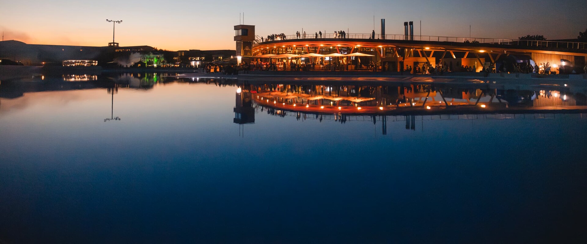 Ob Surfer, Besucher oder Gast im Restaurant oder auf einem Event, die Surftown in München bietet Erlebnisse, an die man sich erinnert. Das Bild zeigt eine Aufnahme des Geländes bei Dämmerung, das Wasser der Surfanlage liegt spiegelglatt da und wirft die Reflexion des beleuchteten Gebäudekomplexes mit Restaurant und Walkway über die Rooftop-Terrace. An der Brüstung sieht man die Silhouetten von Besuchern, die die Aussicht genießen.